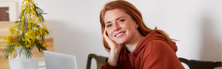 Kvinne som smiler og lener seg på hånden med albuen på bordet og har en pc foran seg 